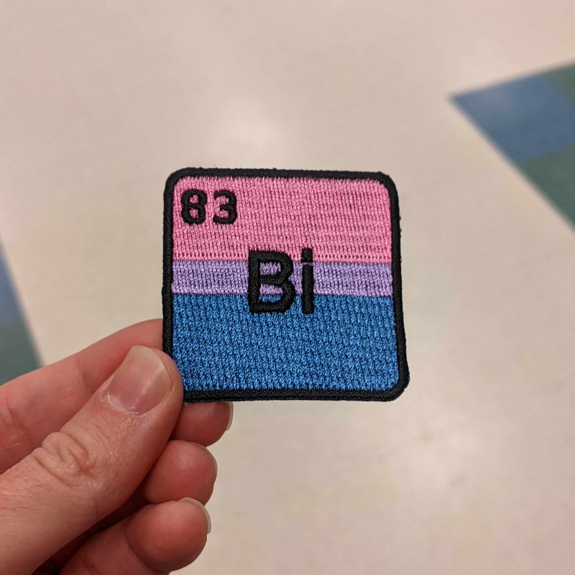 An iron-on patch featuring the chemical symbol for Bismuth with pink and blue coloring, held between fingers.