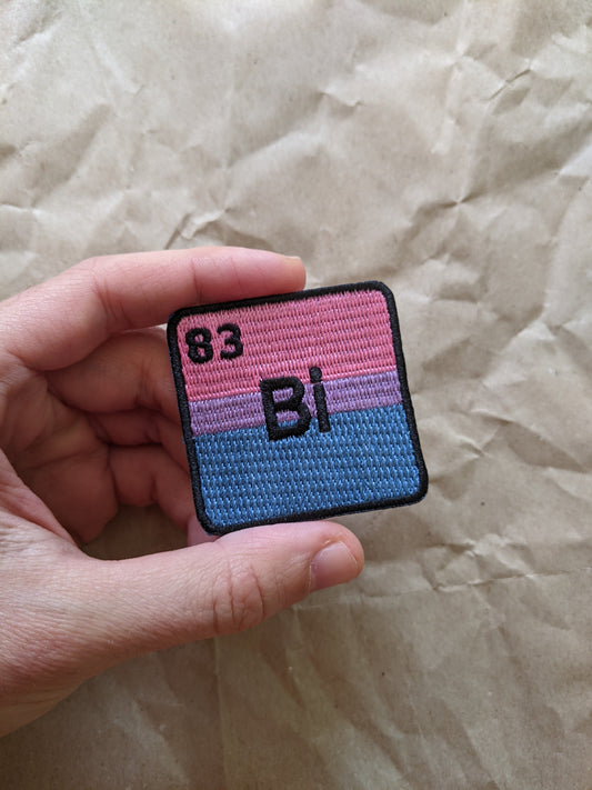 Hand holding a square patch with pink and blue design and '83 BI' text on a textured beige background